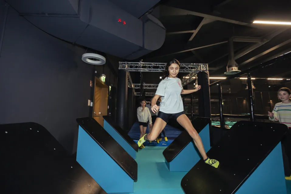Kind balanciert über Hindernisse auf einem Ninja-Parcours in einer Indoor-Trampolinhalle mit Action- und Geschicklichkeitselementen.